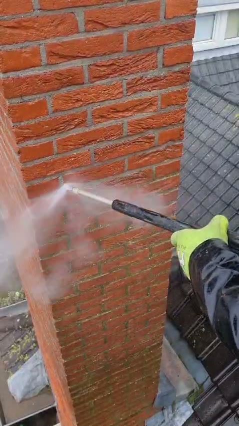 Een hand in een geelgroene werkhandschoen houdt een stoomlans tegen een oranjerode gemetselde schoorsteen; boven is de steen al schoon, onder nog groengrauw