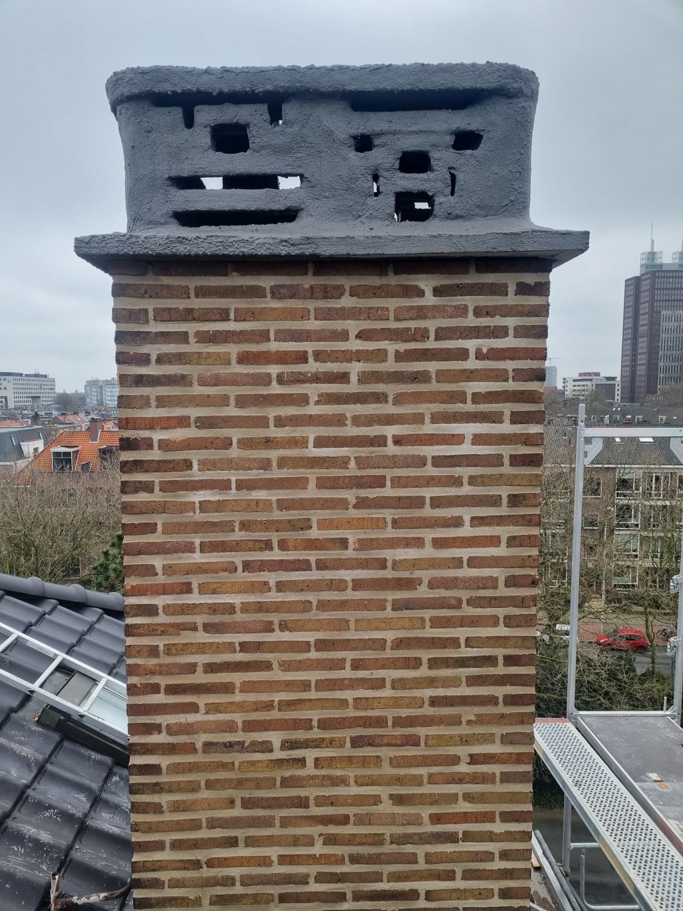Een gemetselde schoorsteen van gele en bruine baksteen met strakke voegen onder een grijze kap met ventilatiegaten; rechts de steiger, op de achtergrond woonblokken en een hoge toren