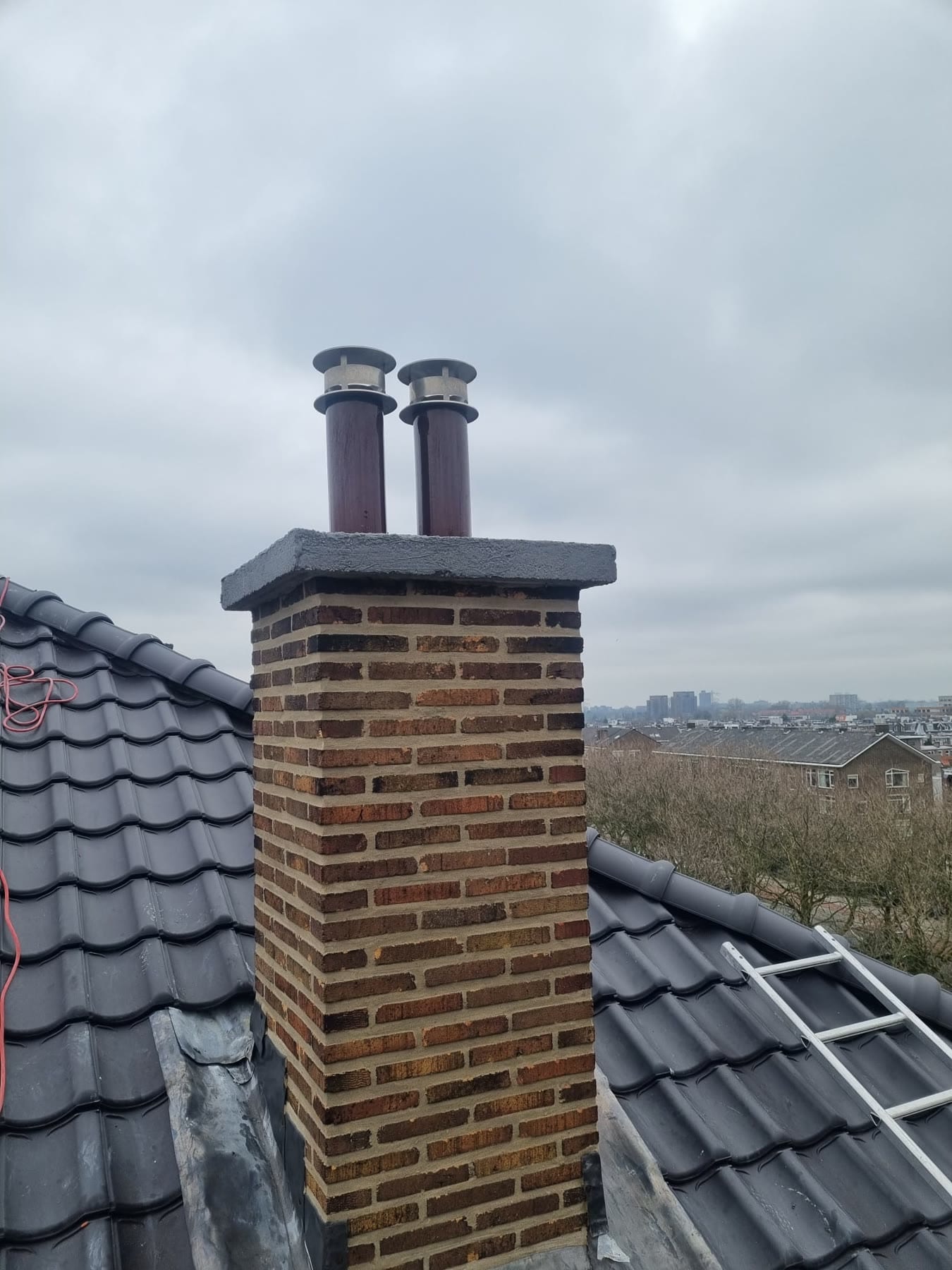 De tweede schoorsteen op hetzelfde dak: gele en bruine baksteen met strakke voegen, een grijze afdekplaat en twee bruine rookkanalen met rvs-kapjes