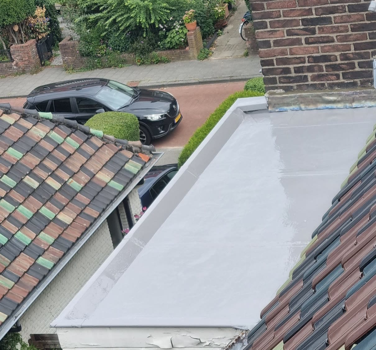 Een plat dak met een gladde, glanzend lichtgrijze coating tot aan de rand, tussen twee pannendaken; beneden de straat met een geparkeerde auto